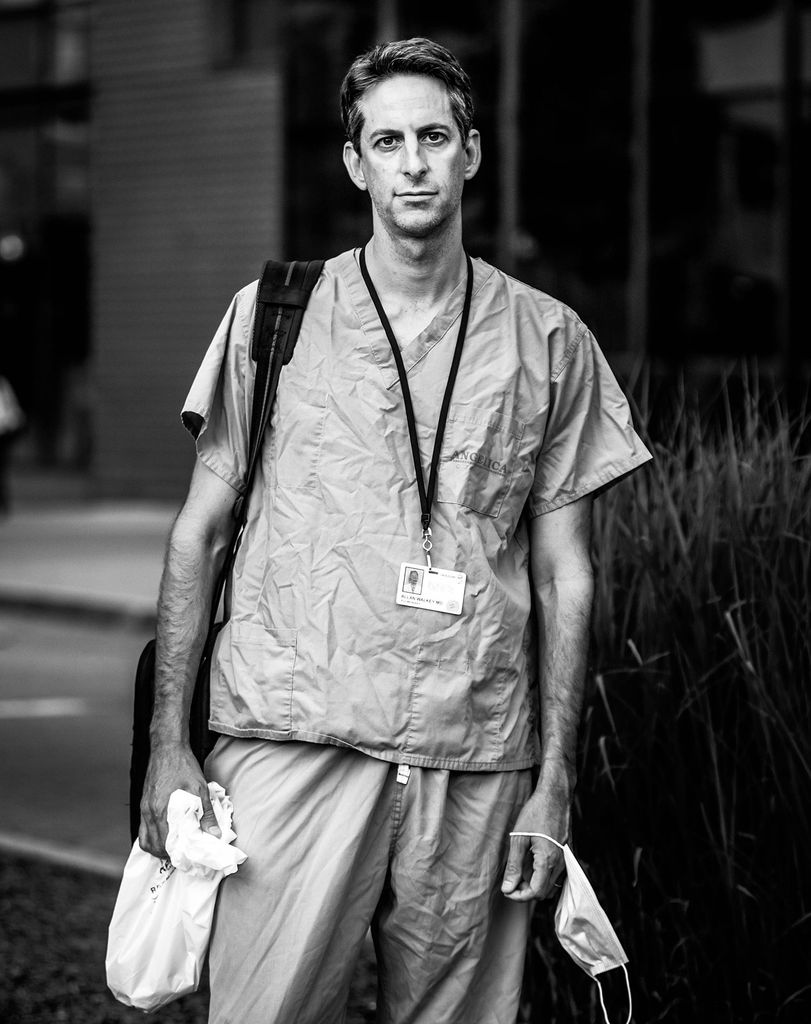 Black and white portrait of Allan Walkey, BMC doctor of critical care medicine, and MED associate professor of medicine. He wears scrubs, a backpack, and holds his facemask in his hand.