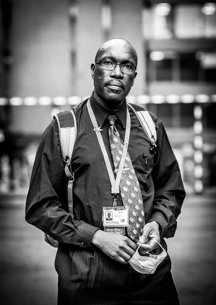 Black and white portrait of Smith Lamothe, BMC patient navigator in a long sleeve button down and tie wearing a backpack.