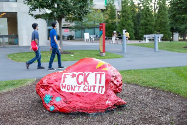 Photo of the BU Rock painted read with a message from BU's student-led public health campaign, "F*CK IT Won't Cut It". In the background, students walk by.