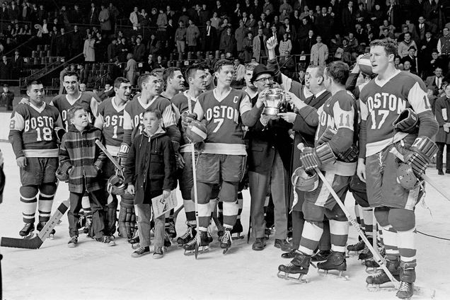 Black and white photo of men's hockey team on the ice after Boston University defeats Northeastern 4-0 in the Beanpot Tournament final game. BU Players include Herb Wakabayashi #18, Jim Quinn #7, Pete McLachlan #11, Bill Hinch #17. David Kelley on ice with his dad, Coach Jack Kelley.