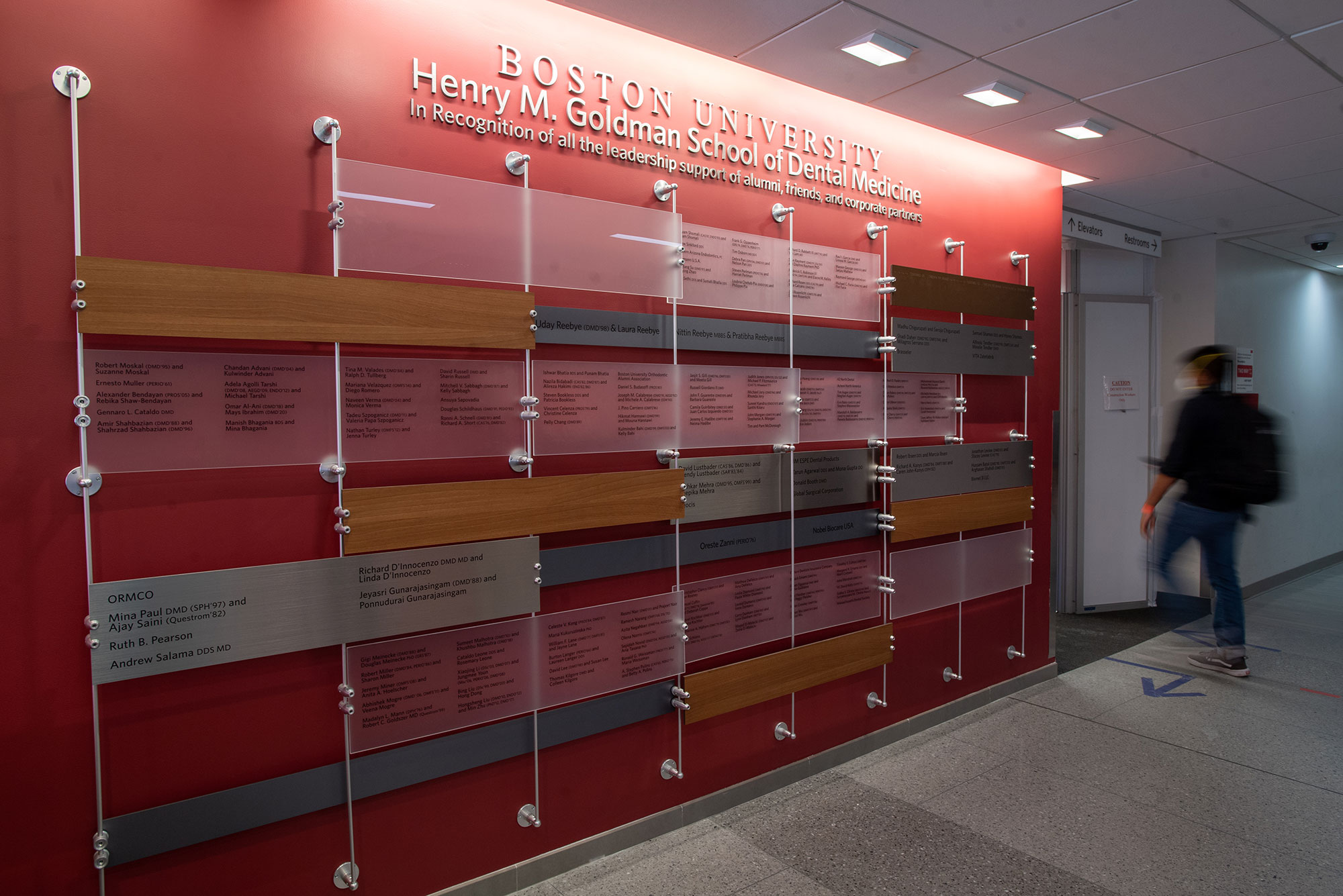 Photo of a reddish orange wall at the Henry M. Goldman School of Dental Medicine covered with plaques that serves as the official donor wall, recognizing all leadership commitments of $25,000 and above made to the school during the Choose to Be Great campaign at Boston University. The plaques' texts are printed on transparent blocks that are hung among silver pipes.