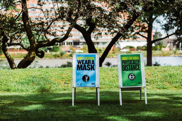 Photo of two signs on BU Beach. One is blue and says “don’t go viral, wear a mask,” while the left one is green and says “don’t go viral, physically distance.” The day is bright and sunny and the charles river is seen in the background.