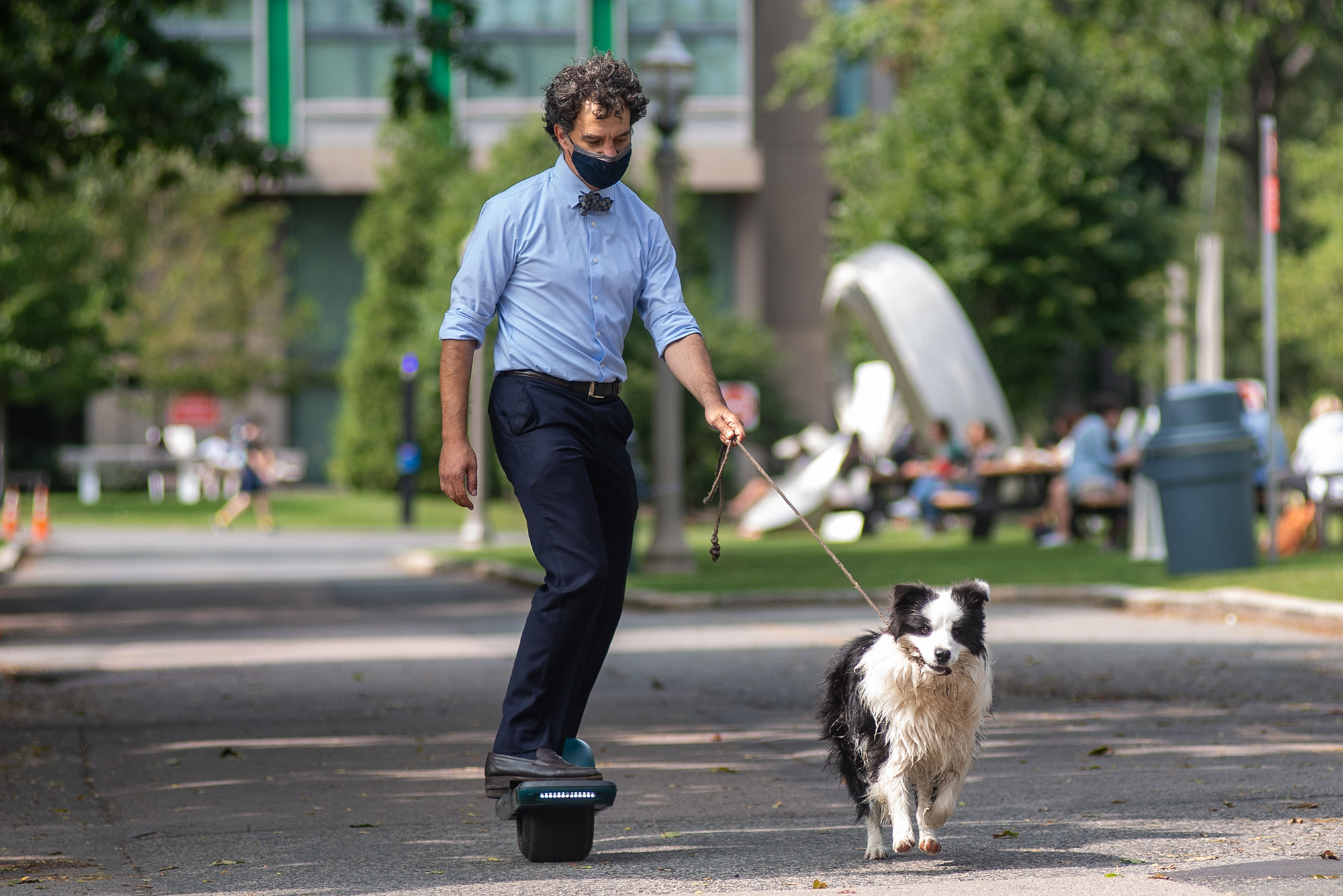 Photo of Professor of earth and environment Bruce Anderson heading back to his office on an electric hoverboard after getting some fresh air with his 1.5 year old pup Juke, a shaggy back and white dog that runs by his side as Anderson holds its leash.