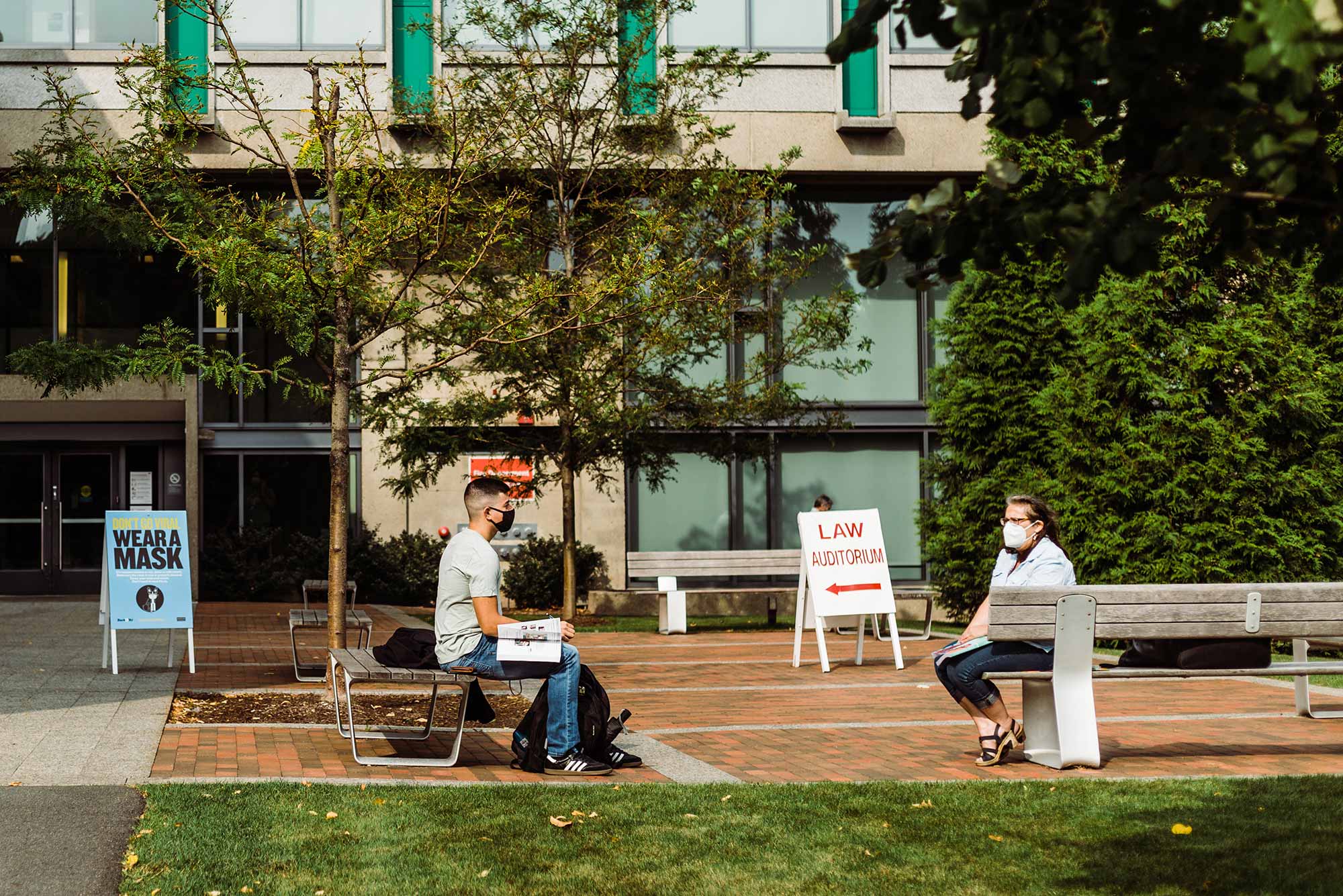 Photo of a student and a faculty/staff member sitting outside the law school and talking at a safe social distance with masks on. A sign behind them says "Law auditorium" with a red arrow pointing to the left. Another blue sign says "Wear a mask."