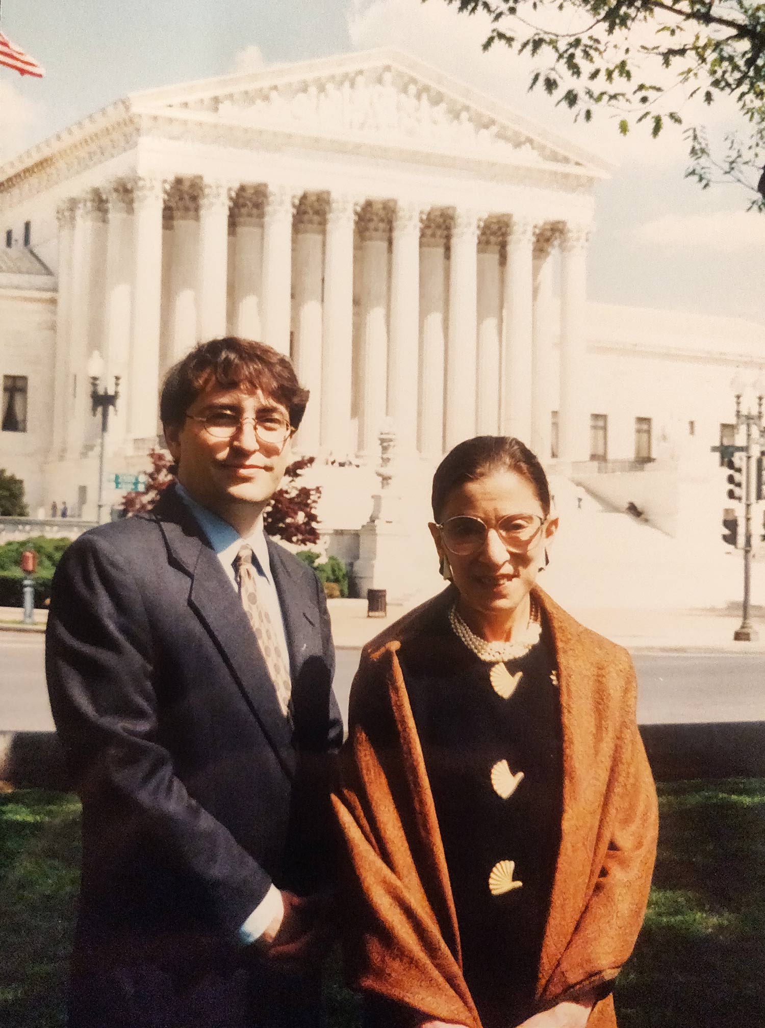 Photo of a printed photo of Jay Wexler (left) with Supreme Court Justice Ruth Bader Ginsburg in 1999, when he was one of her four clerks. Behind them is the facade of the Supreme Court. RBG wears a mustard colored shawl and Wexler wears a suit and tie.