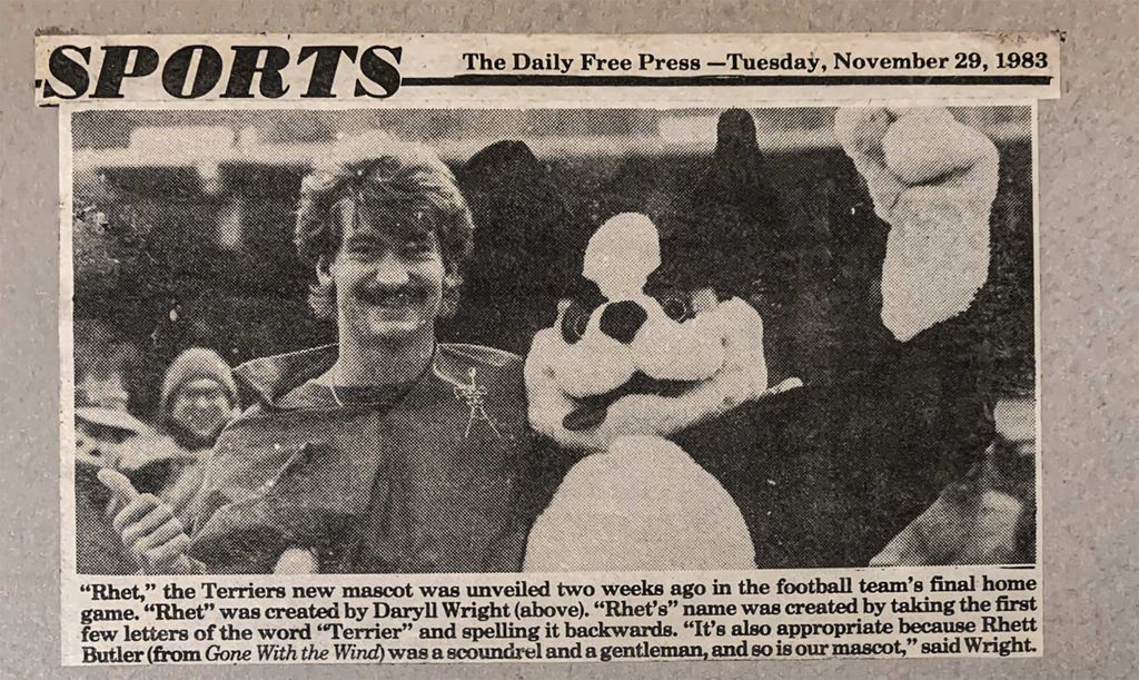 Photo of a newspaper clipping sjpwomg Braden Wright (COM’86), who named “Rhet,” with the mascot at a football game in 1983, before the the second T was added. A headline reads "Sports: The Daily Free Press—Tuesday Tuesday, November 29, 1983.” Caption below reads “Rhet,” the Terriers new mascot was unveiled two weeks ago in the football team’s final home game. “Rhet” was created by Daryll Wright (above). “Rhet’s name was created by taking the first few letters of the word ‘terrier’ and spelling it backwards. “It’s also appropriate because Rhett Butler (from Gone with the Wind) was a scoundrel and a gentleman, and so is our mascot,” said Wright.