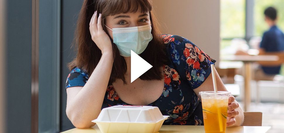 Photo of Brigid Kane (COM’22) sitting at a small dining hall table by herself with a face mask on. A paper to-go container and drink sit in front of her; she rests her hand on her face. Overlay has a video play button.