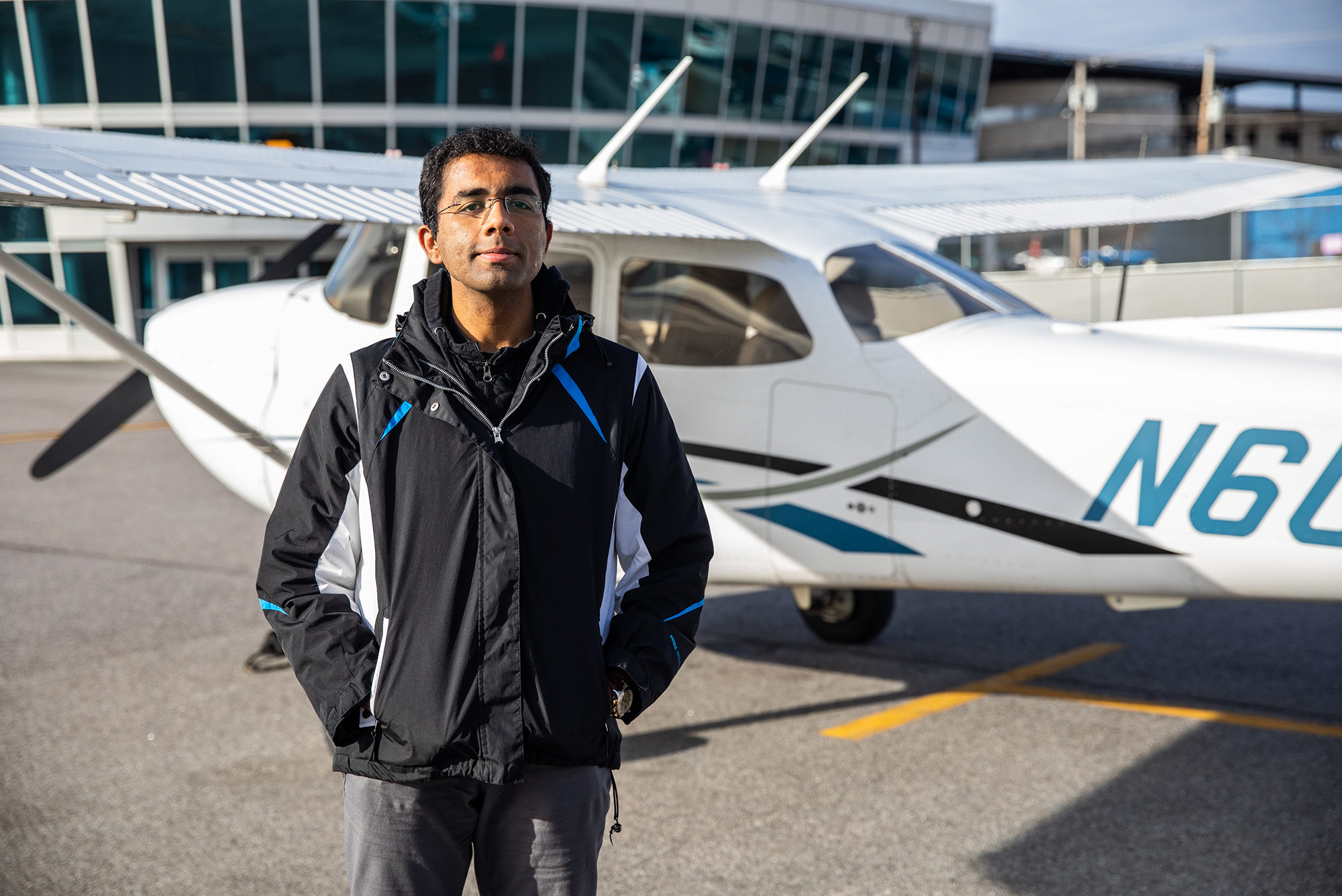 Photo of pilot Dharmesh Tarapore (CAS’18) standing in black jacket in front of a small plane and smiling.