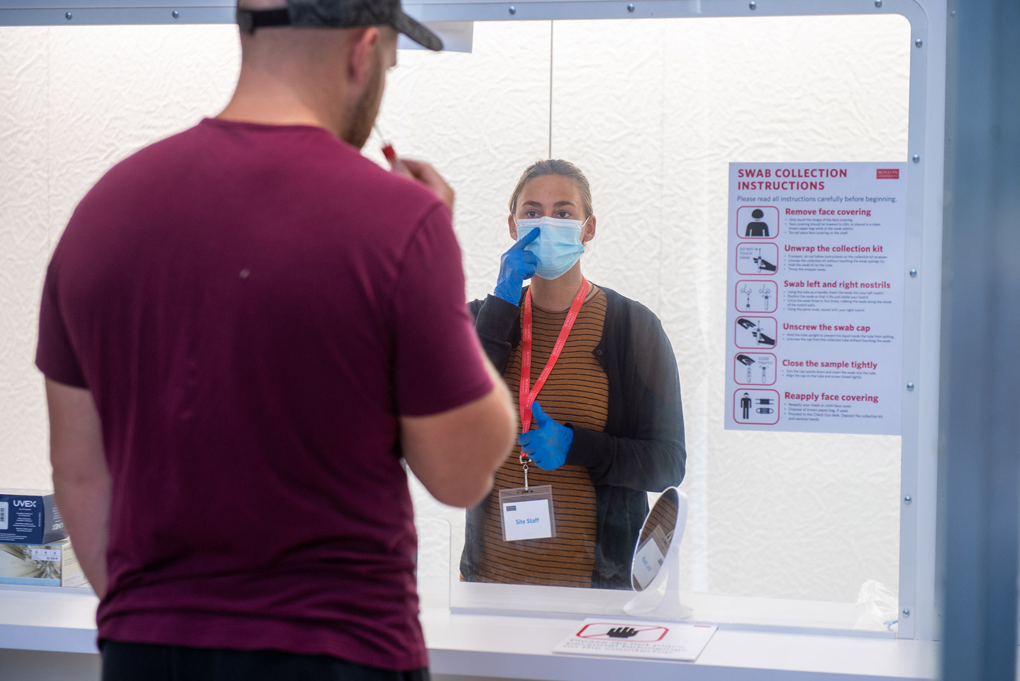 Photo taken at a COVID-19 test collection site. Specialist Jessica Sandberg guides a visitor through the test collection process at the Rajen Kilachand Center August 25, 2020. Sandberg wears a mask and lanyard and stands behind a plastic divider.