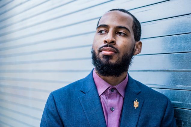 Photo of Jonathan Priester (COM’10) in blue blazer, pink button down, and gold lapel pin. He looks off into the middle distance in front of a corrugated wall.
