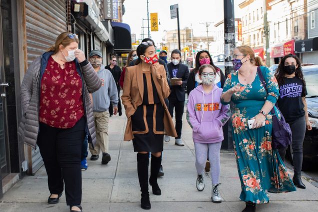 Photo of Alexandria Ocasio-Cortez (CAS’11) (D-N.Y.) campaigning in Queens, N.Y., on October 18, 2020. She wears a red mask and talks to a woman and a young girl to her left.