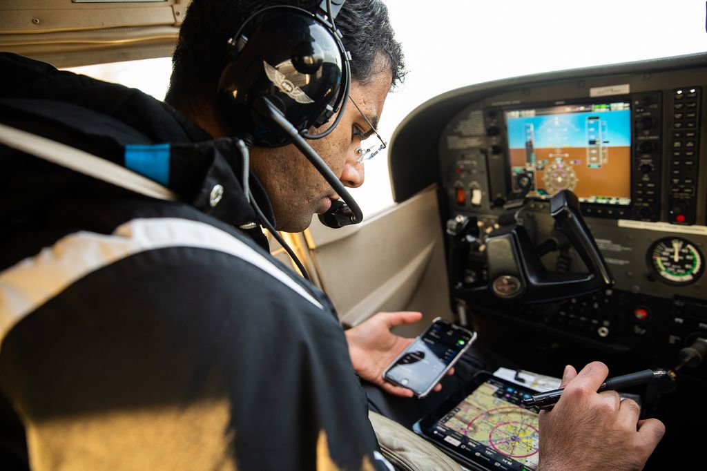 Photo of pilot Dharmesh Tarapore (CAS’18) in the cockpit of a small plane with a headset on. In his lap he balances a phone and a tablet where he monitors other aircrafts in the areas.