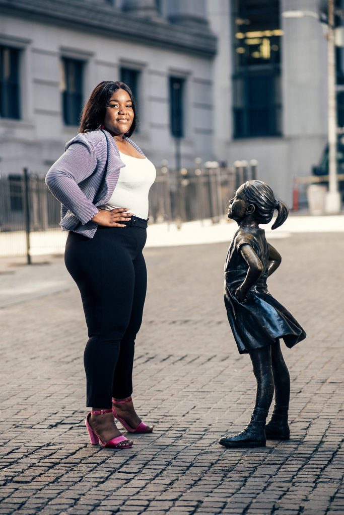 Photo of Delice Nsubayi in a purple blazer and pink heels with her hands on her hips in front of a statue of a young girl.