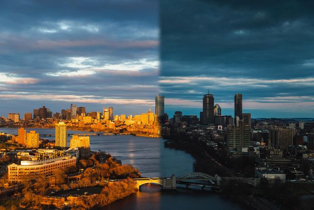 Edited photograph of Boston city skyline taken from the top of a tall building. On the left side, it is morning and the sun is lighting up the buildings. On the right side, the sky is dark and the city lights are on.