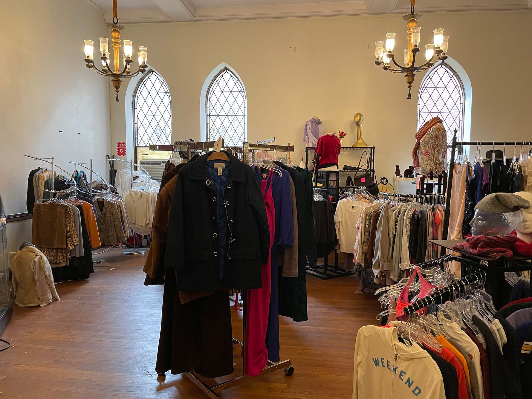 Photo of Thrifty Threads. A thrift shop shows various clothes on rails and in bags around wooden floors. Church style windows line the walls.