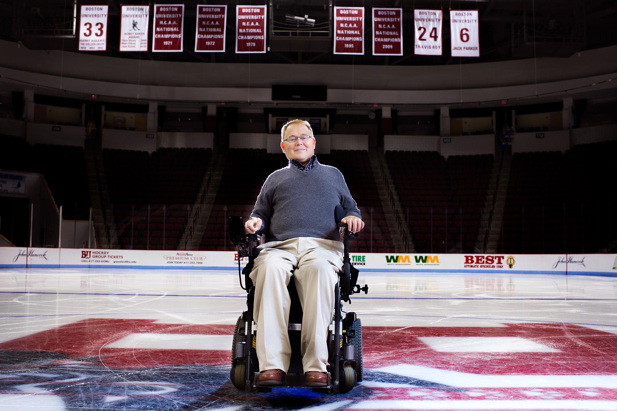 Photo of Travis Roy on Monday, October 6, 2015 at center ice of the Agganis Arena. He wears a grey sweater and his retired jersey number, 24, is seen hanging from the ceiling.