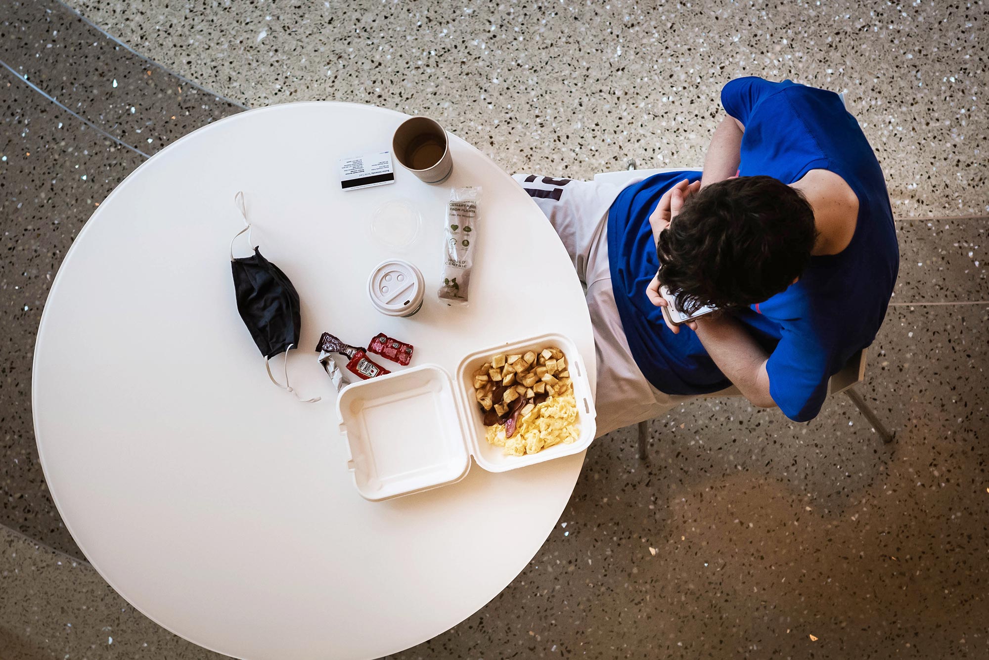 Photo of a student in a blue shirt eating breakfast alone. Their mask and food sits on the table as they look at their phone.