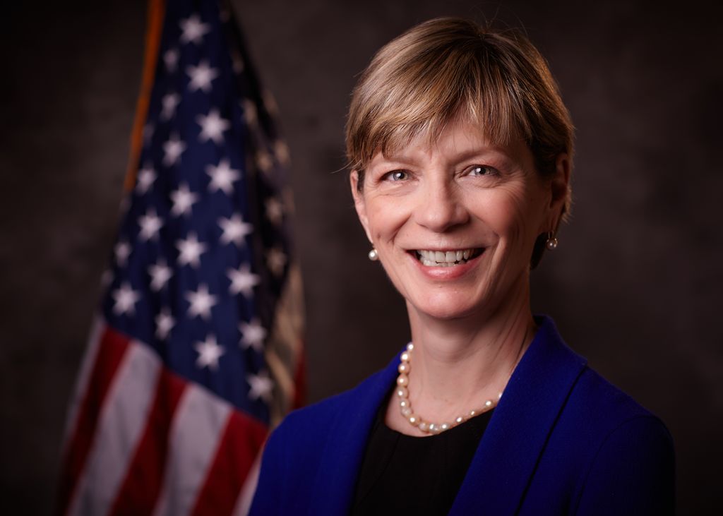 Portrait of Marylou Sudders in pearls and a navy blazer in front of an American flag.