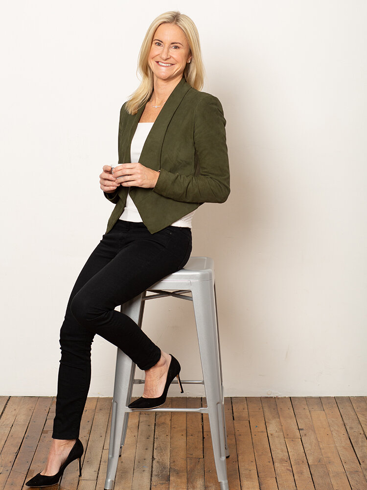 Portrait of Lori Cashman leaning against a gray stole in a green blazer and black high heels.