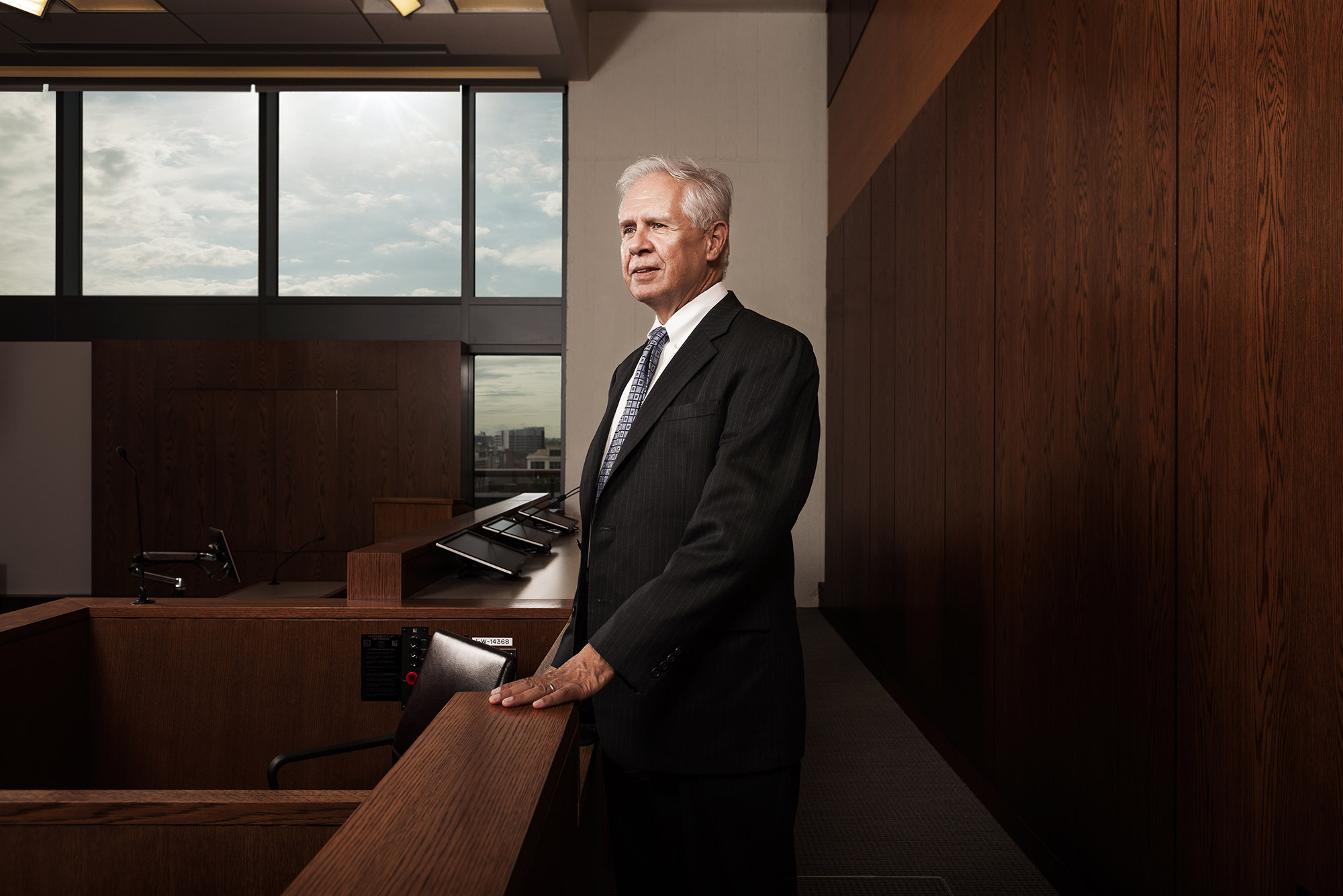 Photo of Robert Burdick (LAW’72), a School of Law clinical professor who directed the Civil Litigation & Justice Program for more than 40 years, standing in a suit, looking off into the middle distance. Bright blue skies are seen in a window behind him.