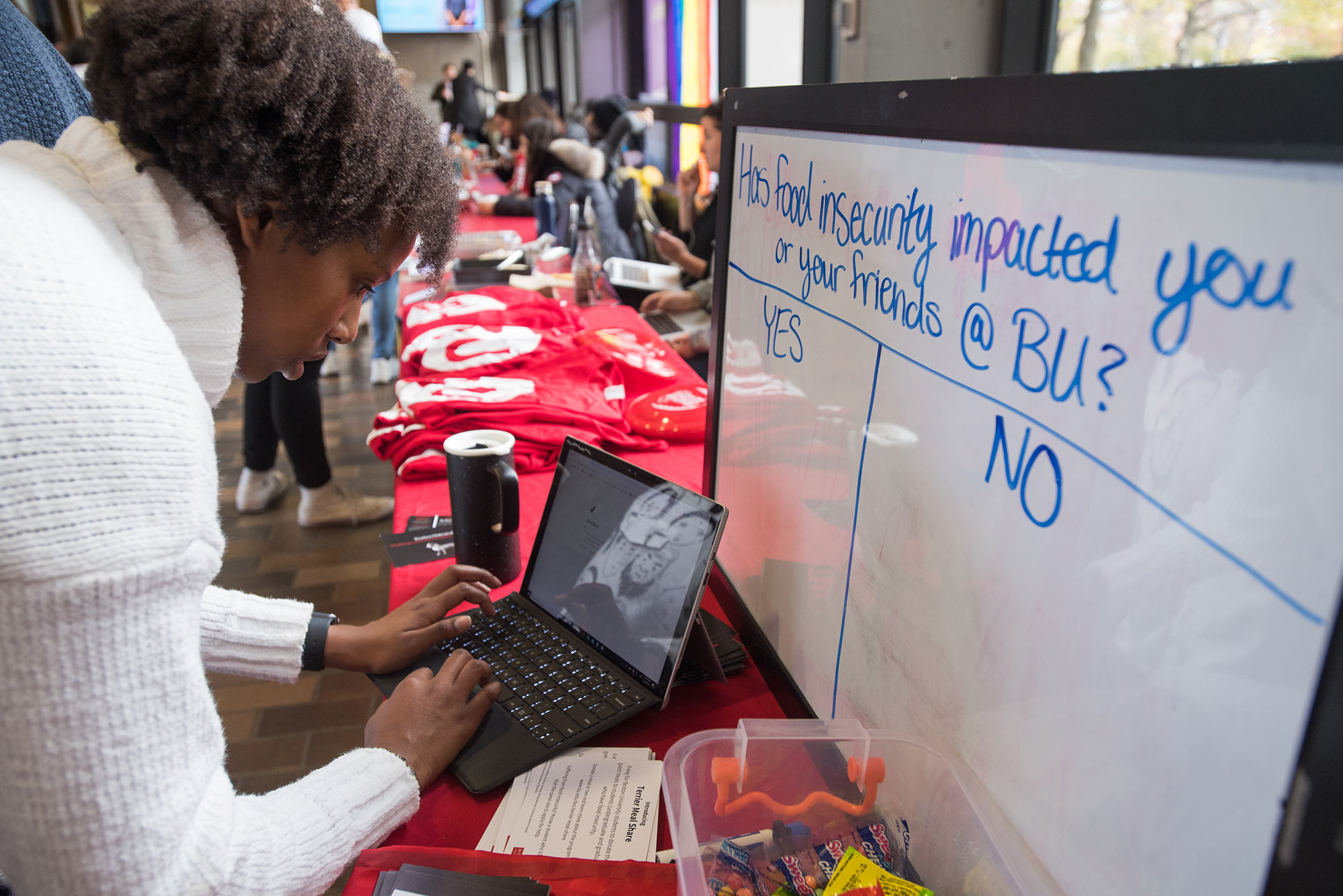 Photo of Michelle Njoroge (CAS,SAR'21), Director of the Department of Social Advocacy for the Student Government during a tabling event in which the group got the word out about Terrier Meal Sharing. Mjoroge sets-up the sign-up system on a laptop at a booth, a white board on the table reads “Has food insecurity impacted you or your friends at BU? Yes or No” with columns for responses.