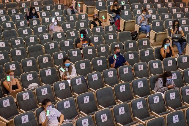 Photo of a group of students sitting in an auditorium as they show their cleared confirmation status on their cellphones at the start of Professor Elizabeth Co’s Human Physiology (BI 211) class in Morse Auditorium on the first day of classes September 2. Stickers are placed in chairs throughout the auditorium to help with distancing.