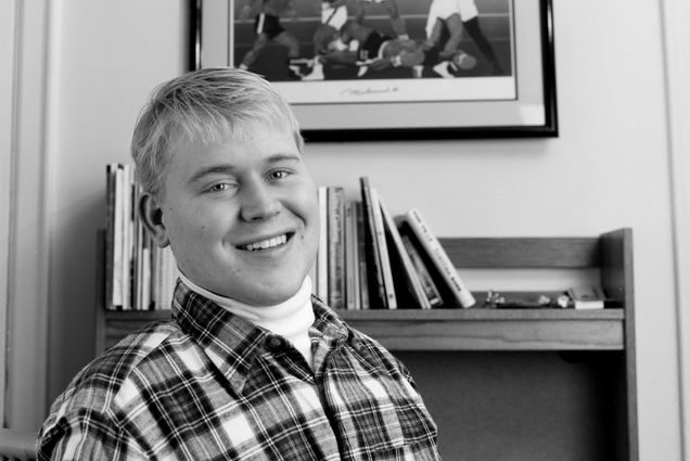 Black and white photo of Roy in his dorm room in 1997, two years after his accident. He wears a white turtle neck and flannel shirt and smiles wide. A photo of Muhammad Ali boxing is behind him above a book shelf.