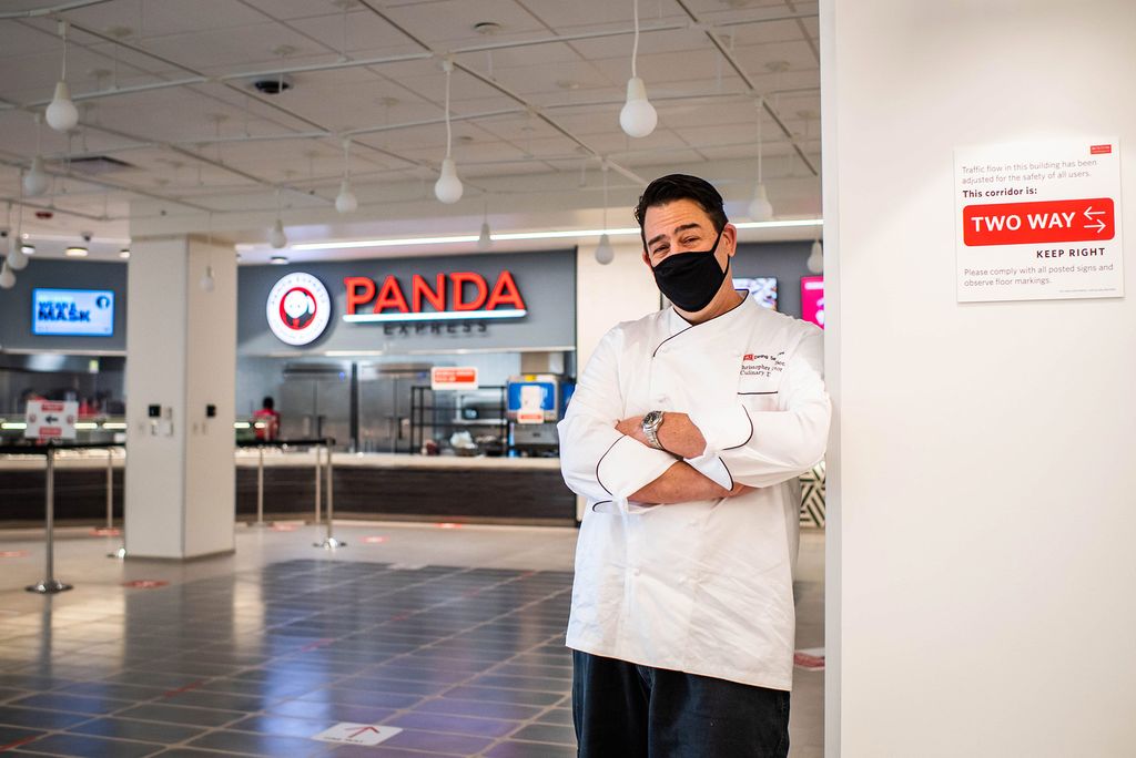 Photo du directeur culinaire du campus, Christopher Bee, responsable d'un grand nombre de saveurs et d'offres que l'on trouve dans tout le Food Hall rénové de GSU, posant les bras croisés devant le favori de la communauté, Panda Express. Il se tient dans ses blancs de chef et un masque noir.