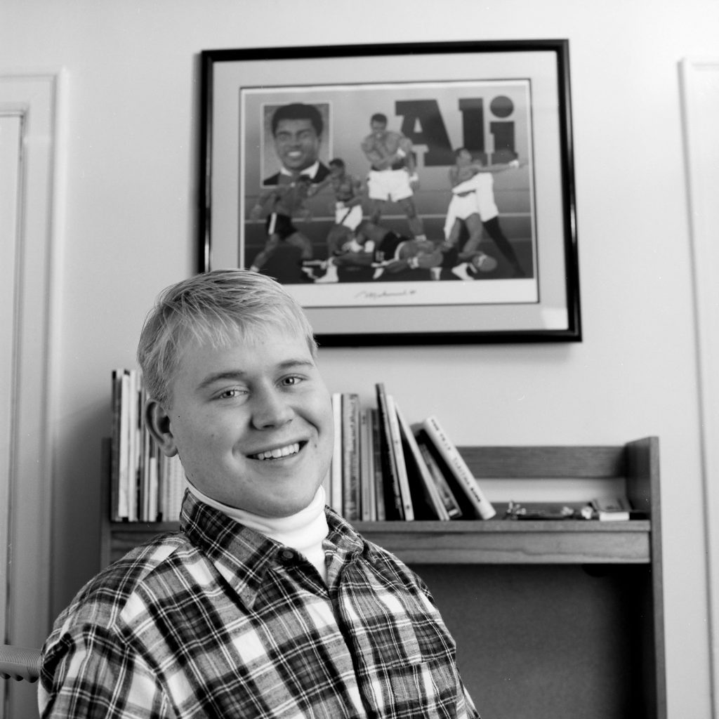 Black and white photo of Roy in his dorm room in 1997, two years after his accident. He wears a white turtle neck and flannel shirt and smiles wide. A photo of Muhammad Ali boxing is behind him above a book shelf.