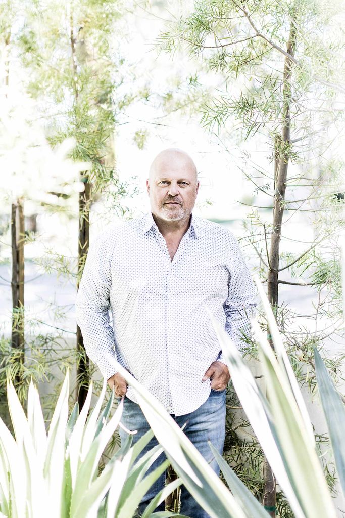 Portrait of actor Michael Chiklis (’85), standing in a white button down with blue dots amongst some pine trees and succulents.