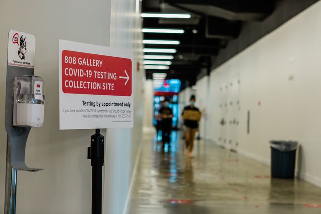 Photo from inside the Covid-19 collection site at 808 Commonwealth Ave taken in August, 2020. A sign reads “808 Gallery COVID-19 Testing Collection Site” with a red arrow, below it reads “testing by appointment only.” A hand sanitizing station is next to the sign, it has a photo of Rhett above it.