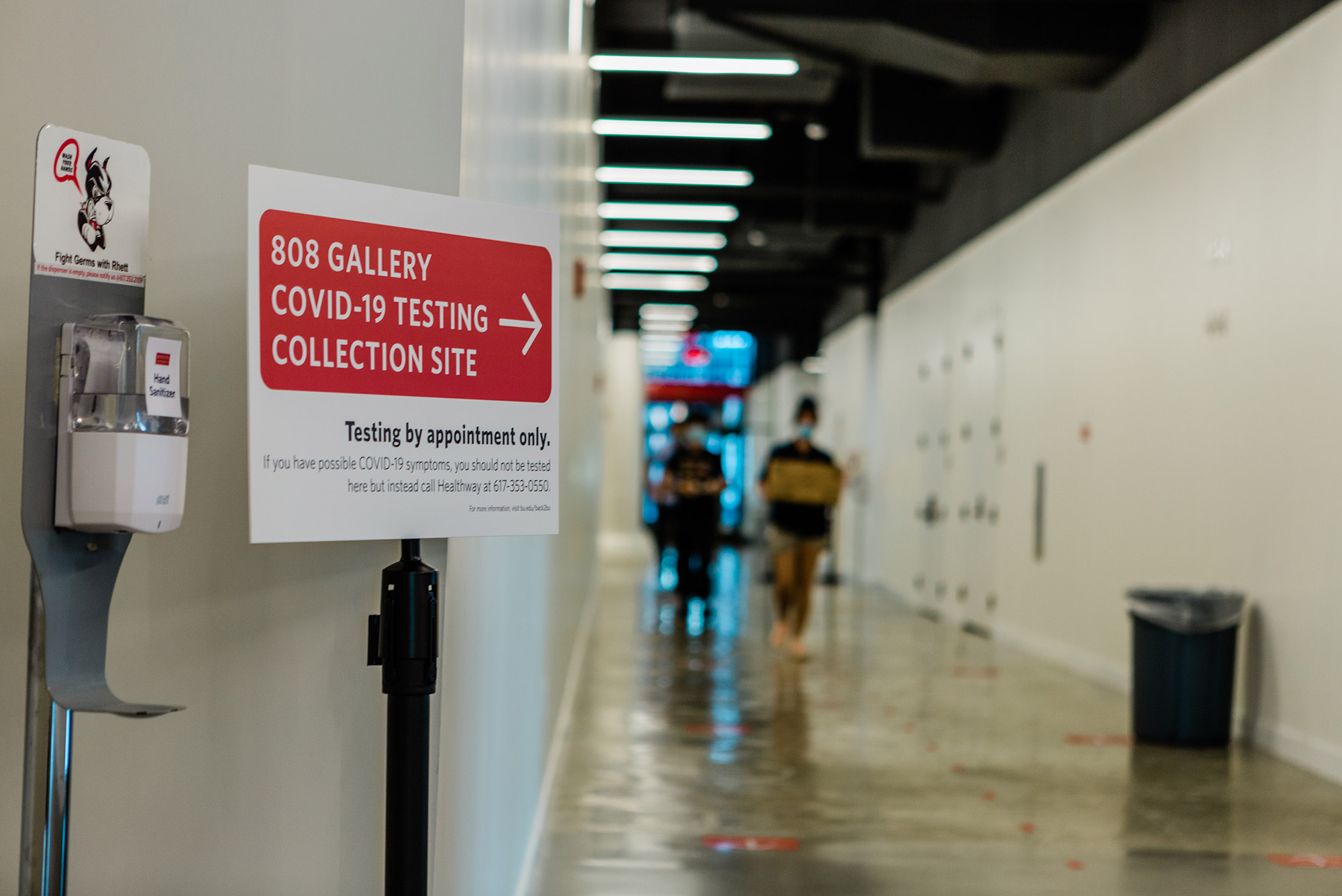 Photo from inside the Covid-19 collection site at 808 Commonwealth Ave taken in August, 2020. A sign reads “808 Gallery COVID-19 Testing Collection Site” with a red arrow, below it reads “testing by appointment only.” A hand sanitizing station is next to the sign, it has a photo of Rhett above it.