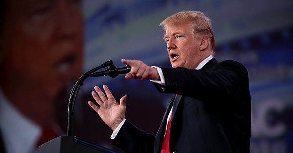 Photo of President of the United States Donald Trump speaking at the 2018 Conservative Political Action Conference (CPAC) in National Harbor, Maryland. He stands at a podium in front of a big screen and points at the crowd. He has a suit and red tie on.