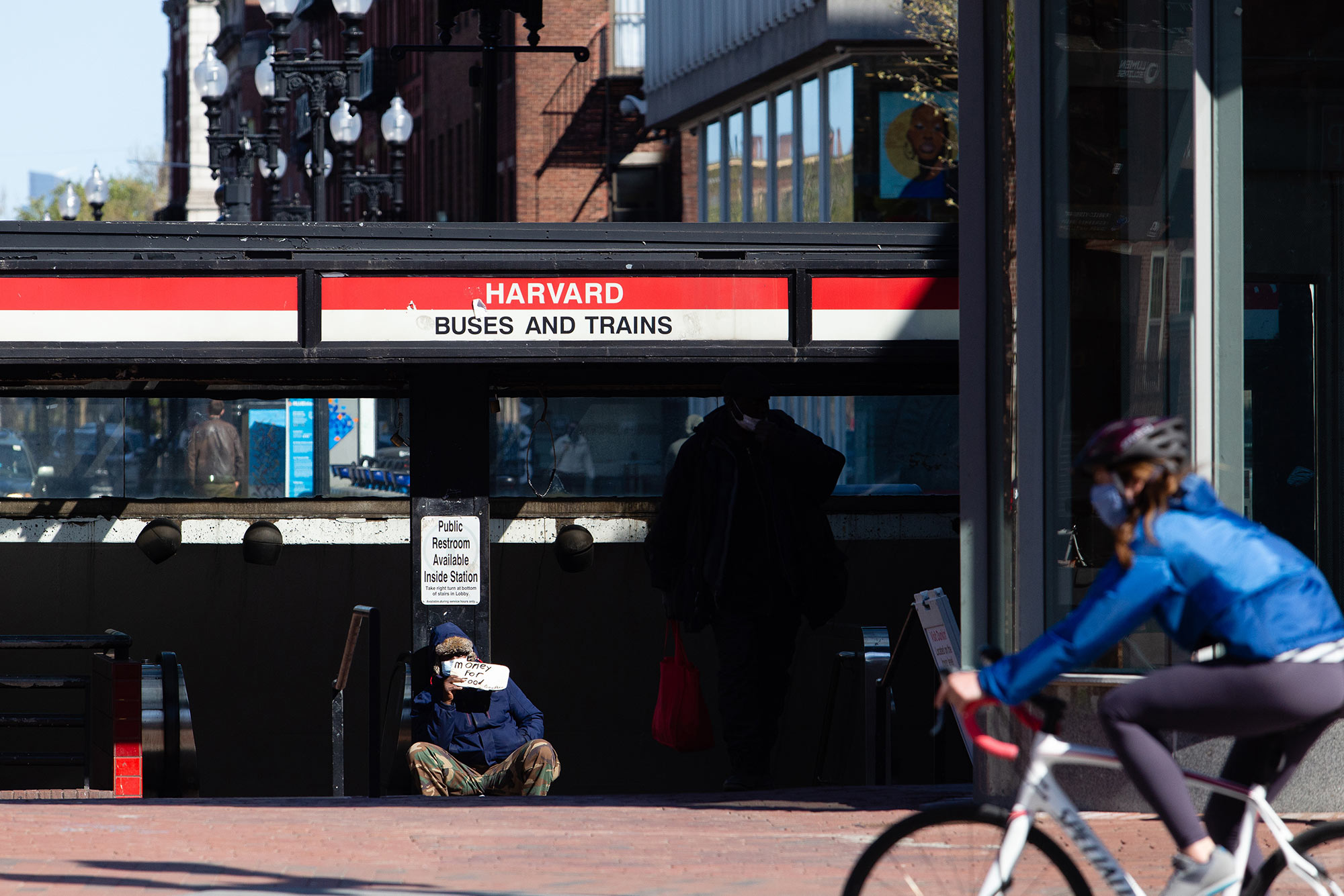 A man pan handles outside the Harvard MBTA Station on May 5, 2020. The man holds a small sign that says "money for food." In the foreground, a woman in a blue jacket rides by on a bicycle.