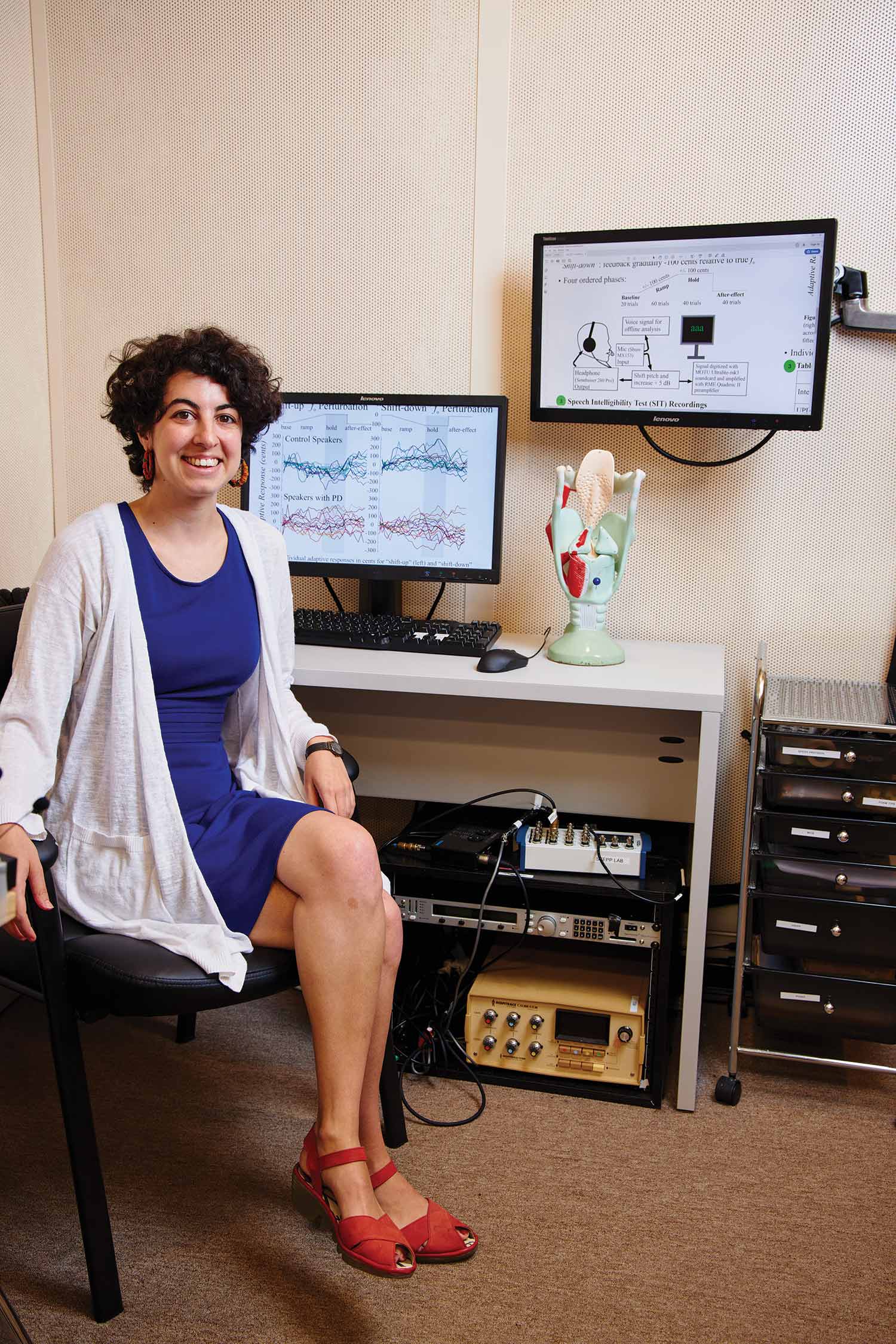 Photo of Defne Abur in a white sweater and bright blue dress and red shoes sitting at her desk in front of two computer screens. On her desk is a medical model of the lungs.