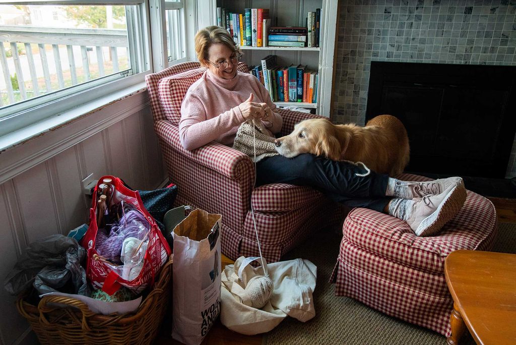 Photo of Megan Nocivelli, lecturer, administrative sciences, Metropolitan College, sitting in her home smiling with her dog on her lap as she knits. Yarn is seen in bags on the ground around her chair.