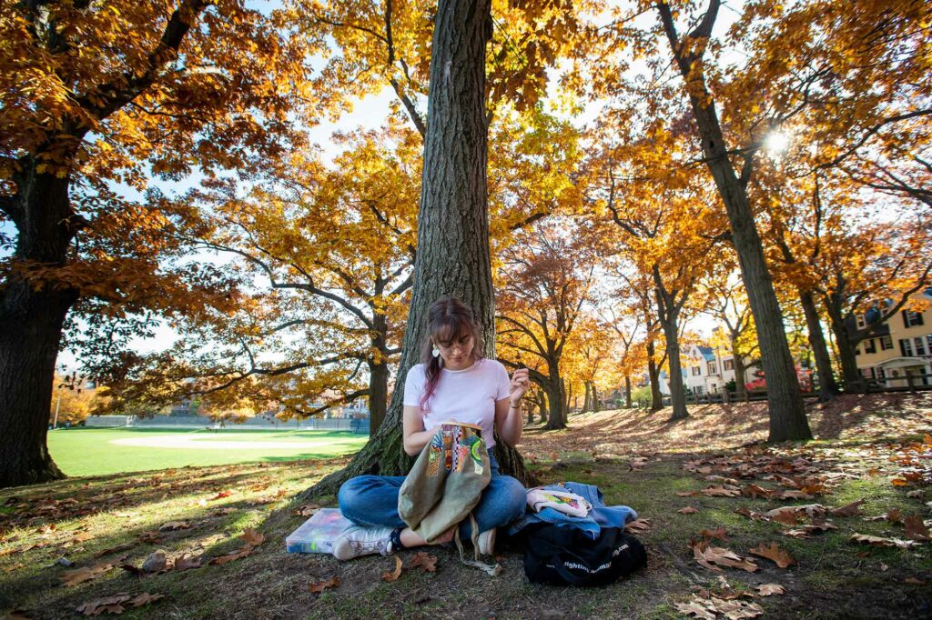 Photo of Sarah Kula (LAW’21) sitting at the base of a tree with golden, fall-colored leaves while working on an embroidery project. 