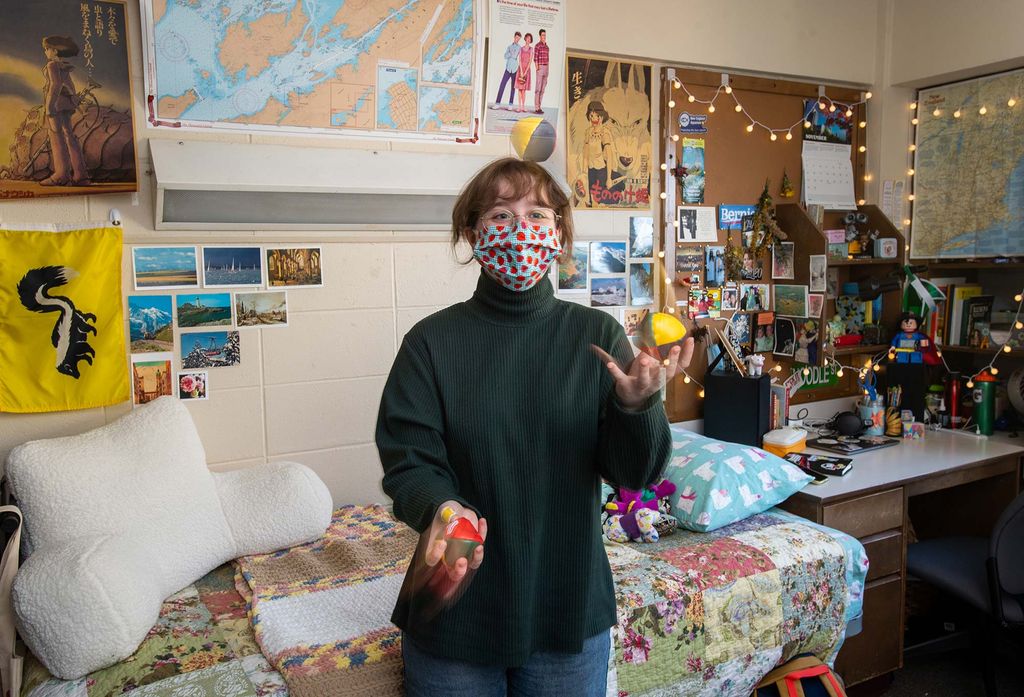 Emma French in her Warren Towers dorm room practicing her juggling. She wears a spotted face mask; her dorm walls are covered with maps, photos, and cork boards.