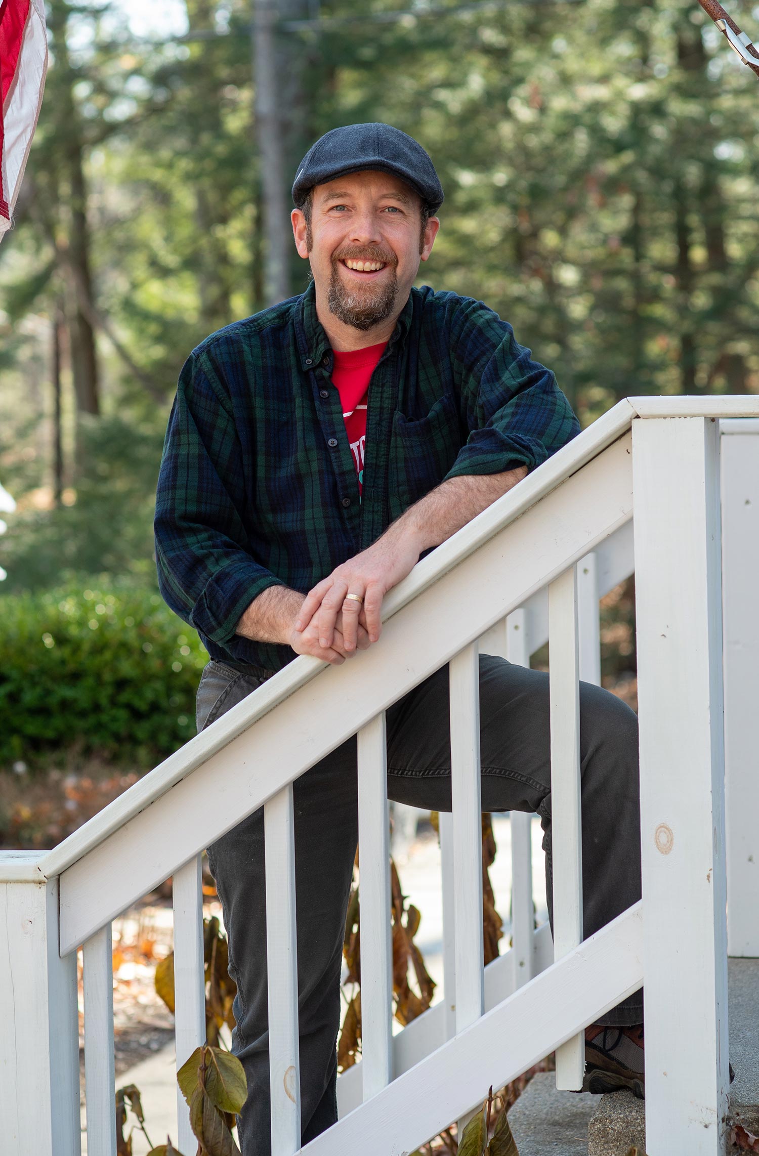 Photo of Paul Hutchinson smiling as he leans on a white wooden railing. He wears a flannel and a hat.