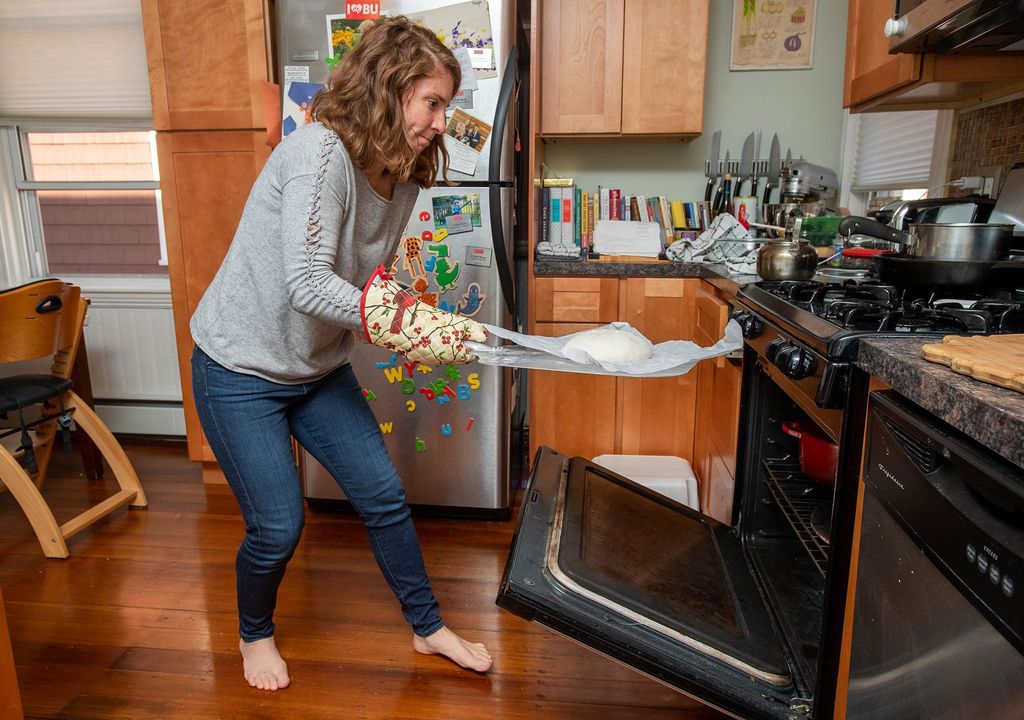 Photo of Katherine Meyer Moran with one of her loaves of sourdough bread at her home in Brookline. The oven is open as she quickly goes to slide in the bread. A fridge with fridge magnets are seen in the background.