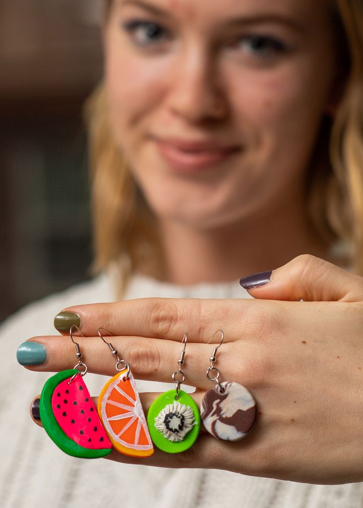 Grace shows off some of her playful earrings, a watermelon, orange, kiwi, earrings hang from her hand.