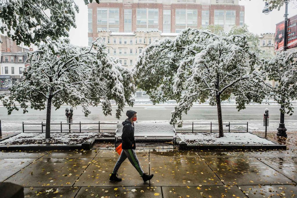 Photo of a late autumn snow storm in October 30 that dropped four inches of snow in Boston. A person in a hat and red fade mask walks down Comm Ave as snow falls.