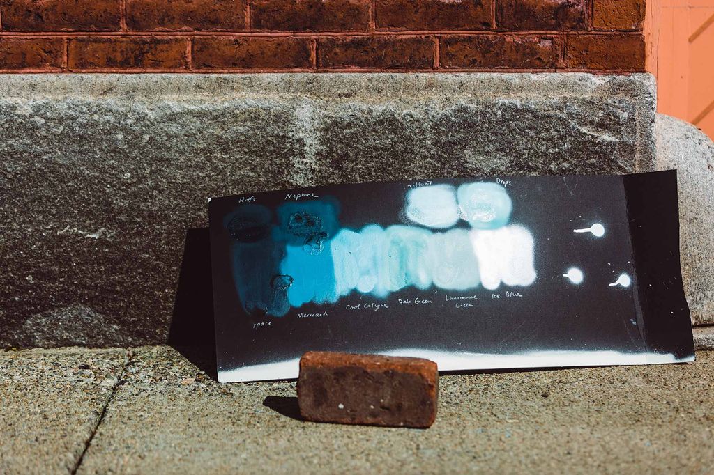 Photo of a piece of wood painted black behind a brick on a sidewalk. The black board is painted with streaks of different shades of blue.