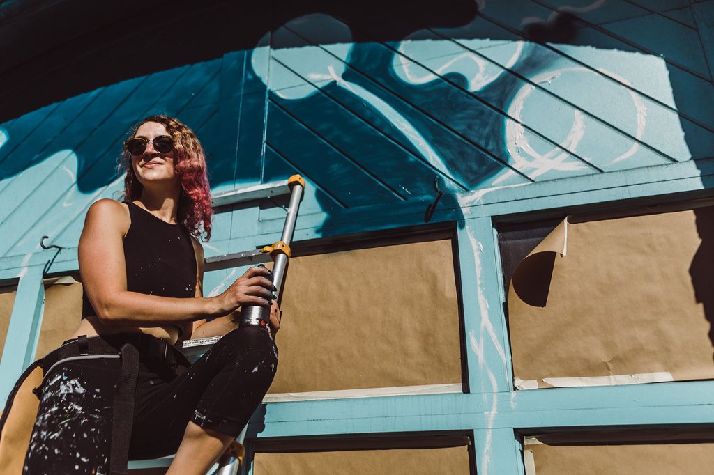 Photo of muralist Lena McCarthy from the front as she stands on a ladder in sunglasses and tool belt with a spray paint can in her hand; she wears all black but some of her clothes are covered with paint. She looks contently out away from the wall and smiles.