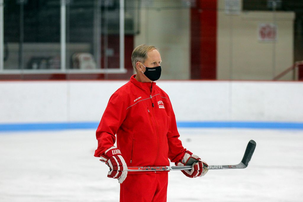 Photo of Brian Durocher, who has been a BU Women’s hockey coach for 16 years, in a black face mask, hockey gloves, and a red track suit on the ice with a hockey stick in his hands.