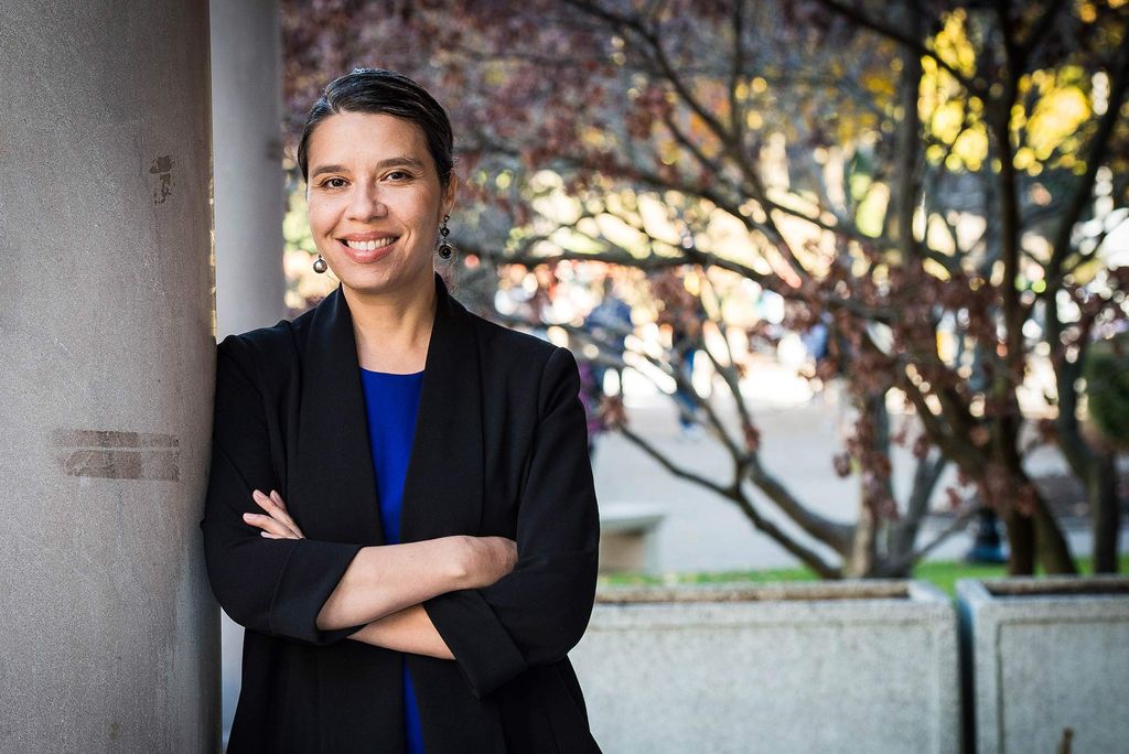 Portrait of Jasmine Gonzales Rose in a black blazer and bright blue blouse with her arms crossed as she smiles, leaning against a cement post. Behind her students on campus are seen. 