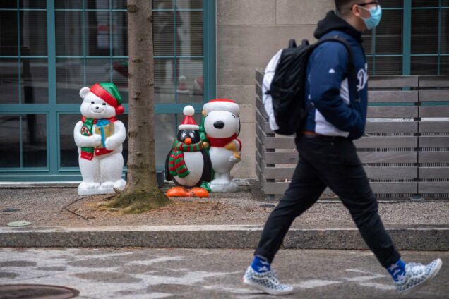 Photo of a three small light-up decorations, a white bear in a red and green hat, a penguin in a Santa hat, and a Snoopy with a Santa hat, at the base of a building. A person in a mask and with a back pack on walks by on the right.