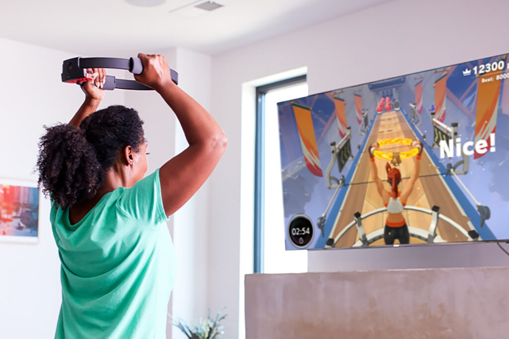 Photo of a woman working out while the Nintendo Switch game Ring Fit Adventure plays on the TV. The TV screen shows an avatar with the ring and words ‘Nice!’