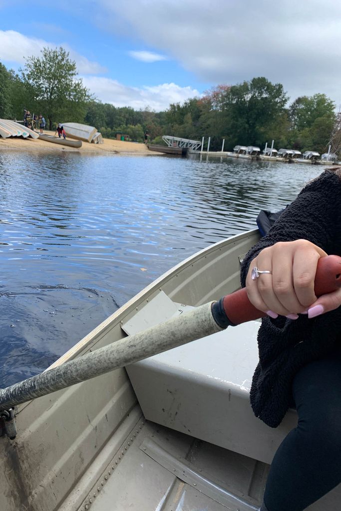 Photo of a hand with pink nail polish and a silver ring rowing a small row boat on a pond. In the background, the pond's beach and a line of row boats is seen.