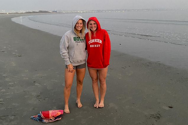 Photo of Jenna Seibold (left) and Sammy Davis (right) who stand barefoot at the shoreline of a beach in sweatshirts, Davis wears a red BU sweatshirt and Seibold wears a gray Endicott sweatshirt. The two look a bit cold after having done a polar plunge.